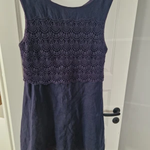 Mörk blå klänning från Flash Woman - Supersnygg mörk blå klänning från Flash Woman i 100% linne. Överdelen har ett detaljerat spetsmönster som ger en unik look. Klänningen är ärmlös och har en rak, enkel passform. Perfekt för dig som gillar stilrena men ändå speciella plagg.