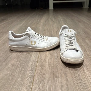 Vita sneakers från Fred Perry - Snygga vita sneakers från Fred Perry med klassisk design. Skorna har guldiga Fred Perry-loggor på sidan, tungan och hälen samt perforerade detaljer. Tillverkade i skinn med vita snören och platt sula. Perfekta för en clean och stilren look.