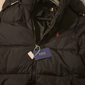 Svart pufferjacka från Polo Ralph Lauren - Svart pufferjacka från Polo Ralph Lauren, huva och dragkedja framtill. Jackan har en röd broderad logga på bröstet och flera praktiska fickor med dragkedja. Perfekt för kalla dagar när du vill hålla stilen.