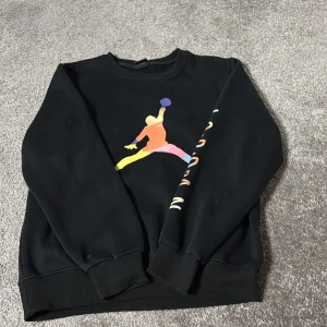Svart Jordan sweatshirt med färgglad logga - Svart sweatshirt från Jordan med en stor färgglad Jumpman-logga på bröstet och texten 'JORDAN' i olika färger längs ena ärmen. Tröjan har rund hals, ribbade muddar och är långärmad. Perfekt för dig som gillar streetwear och sportig stil.