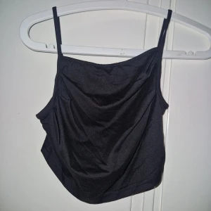 Croppad linnetopp - Säljer en svart croppad linnetopp med tunna axelband och draperad front. Toppen har en enkel och stilren design som passar perfekt till jeans eller kjol. Materialet känns mjukt och följsamt mot huden.