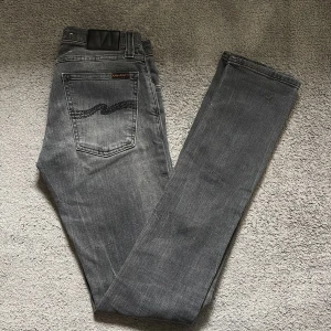 Grå jeans från Nudie Jeans, 27/34 - Snygga grå jeans från Nudie Jeans med klassisk femficksdesign och svart broderad våg på bakfickan. Jeansen har en rak passform och är tillverkade i mjukt denimtyg av bomull. Perfekta för dig som gillar stilrena och tidlösa jeans.