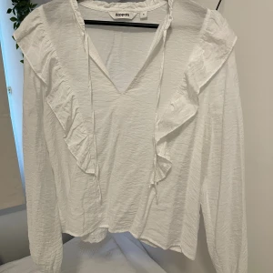 Vit blus med volanger  - Supersöt vit blus. Blusen har långa ärmar, v-ringning med knytband och snygga volangdetaljer framtill och baktill. Materialet är lätt och fint. Passar till det mesta.