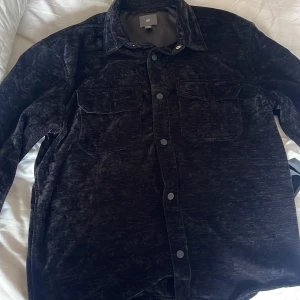 Svart sammetsskjorta från H&M - Mörk, svart skjorta i mjuk sammet från H&M med klassisk krage, tryckknappar och två stora bröstfickor. Skjortan har lång ärm och en loose passform som ger en avslappnad vibe. Perfekt för dig som gillar stilren men ändå unik look.