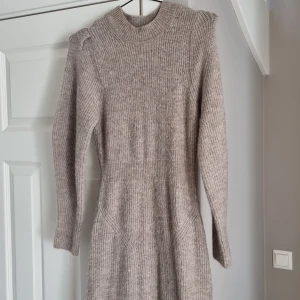 Beige stickad långklänning från Adoore - Mysig beige stickad långklänning från Adoore med ribbad struktur, markerad midja och lång ärm. Klänningen har en liten slits framtill och axeldetaljer som ger extra stilpoäng. Perfekt för dig som vill ha en trendig och bekväm look under kyliga dagar. Använd fåtal ggr.
