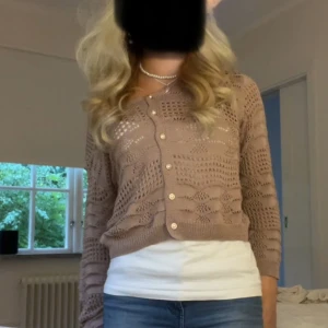 Beige stickad kofta med knappar - Säljer en beige stickad kofta med snyggt hålmönster och pärlemorknappar framtill. Koftan har lång ärm och en croppad passform som ger en trendig look. Perfekt att slänga över en topp till jeans eller kjol.