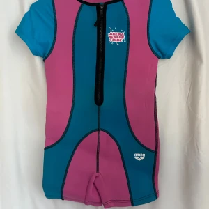 Blå och rosa våt/soldräkt från Arena - Cool våt/soldräkt från Arena Water Tribe i blått och rosa med svarta sömmar och dragkedja framtill. Baddräkten har korta ärmar och ben, tryckt logga och roliga vita figurer på ryggen. Materialet är 80% polyamid och 20% elastan, insidan är 100% neopren.