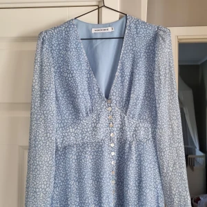 Ljusblå mönstrad klänning från Adoore - Supersöt ljusblå klänning från Adoore med vitt leopardmönster, v-ringning och knappar framtill. Långärmad modell i lätt material, perfekt för vår och sommar. Snygg passform som ger en feminin siluett.