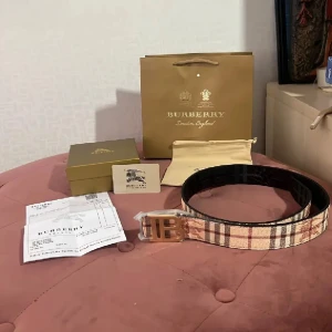 Rutig Burberry bälte med guldspänne - Säljer ett snyggt Burberry bälte med klassiskt rutigt mönster i beige, brun och röd. Bältet har ett stilrent TB-spänne i guld och är tillverkat i skinn med textil på ovansidan. Perfekt accessoar för att lyfta din outfit med lyxig känsla och ikonisk design. Allt som finns på bilden igår storlek 100-40