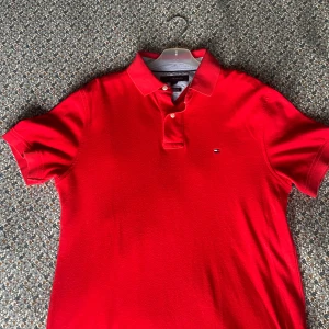 Röd pikétröja från Tommy Hilfiger - Klassisk röd pikétröja från Tommy Hilfiger i custom fit. Tröjan har korta ärmar, krage med två knappar och den ikoniska lilla loggan broderad på bröstet. Perfekt för en clean och sportig stil. Tillverkad i mjuk bomull.