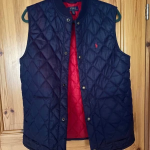 Quiltad blå dunväst från Polo Ralph Lauren - Snygg quiltad dunväst från Polo Ralph Lauren i mörkblått med röd insida. Västen har tryckknappar framtill, ståkrage och Polo-logga broderad på bröstet. Perfekt för lager-på-lager och har en klassisk, sportig look.