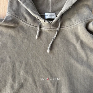 Grå hoodie från Saint Laurent - Säljer en grå hoodie från Saint Laurent med broderad logga på bröstet. Tröjan har huva med snörning, magficka och ribbade muddar. Perfekt för dig som gillar stilrena och exklusiva plagg med streetkänsla.