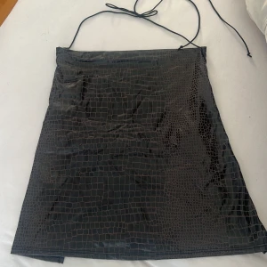 Svart topp med crocomönster - Snygg svart topp med crocoinspirerat mönster och tunna axelband som knyts i nacken. Klänningen har en slät, glansig yta och är figurnära med minimalistisk design. Perfekt för dig som gillar trendiga detaljer och vill sticka ut.