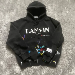 Svart Lanvin hoodie - Svart hoodie från Lanvin Paris med stor vit logga på bröstet och coola färgstänk i blått, rött, gult och lila. Tröjan har huva med snörning och en patch med texten 'Art on Display Courtesy of Josué Thomas' på nedre delen. Perfekt statement-plagg för dig som gillar streetstyle.