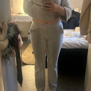 Grå mjukisbyxor från Juicy Couture - Grå mjukisbyxor från Juicy Couture med broderad logga på höften. Byxorna har raka ben, snörning i midjan och fickor både fram och bak med knappdetaljer. Perfekta för en chill och avslappnad stil.