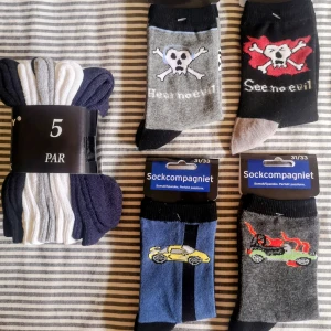 9 Par Pojkstrumpor Stl 31 /34 - Helt nya. Paketet innehåller 5 par i svart, vitt och grått samt fyra par med motiv som dödskallar och bilar, i mjuk bomullsblandning med stretch och passar perfekt för dig som gillar att sticka ut.