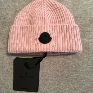 Rosa ribbstickad mössa från Moncler - Snygg rosa ribbstickad mössa från Moncler med svart logopatch framtill. Tillverkad i Italien och gjord i mjukt, lyxigt material som håller dig varm. Nyskick aldrig använd, nypris-2899 mitt pris-1100 pris kan diskuteras vid snabba affärer 