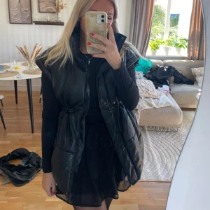 Svart skinnimitation väst LXA, Linn Ahlborg, M - Säljer en svart, väst i syntetmaterial med skinnlook från LXA, Linn Ahlborg . Västen har hög krage, dragsko i midjan för snygg passform och stora fickor framtill. Stängs med dragkedja och tryckknappar. Storlek M. Har fått en liten defekt längst ner vid dragkedjan, se bild 4, inget man tänker på .