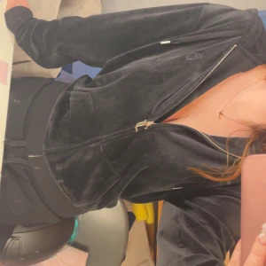 Svart Juicy Couture tröja - Svart velourjacka från Juicy Couture med dragkedja framtill och huva. Jackan har fickor på sidorna och diskret broderad logga på bröstet. Perfekt för en chill och trendig look. Nytt skick