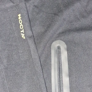 Svart NOCTA x Nike vindjacka med huva - Svart vindjacka från Nike NOCTA med huva och halv dragkedja. Jackan har diskreta guldiga loggor, vattentäta dragkedjor på fickorna och sportig design. Perfekt för dig som gillar streetwear och vill ha en clean look.