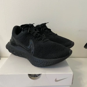 Svarta Nike React Flyknit sneakers - Säljer ett par helsvarta Nike React Flyknit sneakers i storlek 44. Skorna har en stickad ovandel, diskret Nike-logga på sidan och en chunky, dämpad sula för extra komfort. Perfekta för dig som gillar stilrena och sportiga skor med modern känsla.
