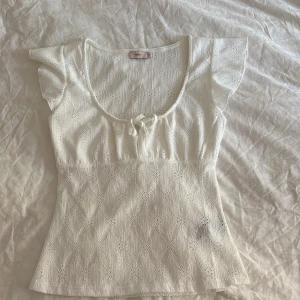 Vit spetsblus med rosett från Nelly - Supersöt vit blus från Nelly med spetsmönster och liten rosett framtill. Blusen har korta volangärmar, rundad urringning och är figurnära i modellen. Perfekt för dig som gillar romantiska detaljer och ljusa färger.