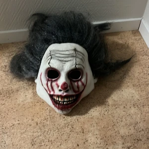 Kuslig clownmask med svart hår - Säljer en skräckinjagande clownmask med vitt ansikte, svarta och röda detaljer samt stort, rufsigt svart hår. Masken har markerade tänder, röda linjer runt ögonen och munnen samt en röd näsa. Tillverkad i syntetmaterial och passar perfekt till halloween eller maskerad.