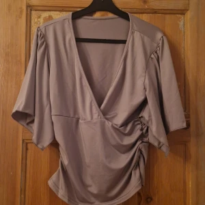 Ljuslila omlottopp med v-ringning - Säljer en ljuslila omlottopp med v-ringning och korta, vida ärmar. Toppen har snygg rynkdetalj på sidan som ger en cool draperad effekt. Perfekt för dig som gillar en avslappnad men ändå trendig look.