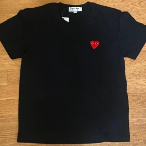 Svart t-shirt Comme des Garçons Play - Svart Ami Paris t-shirt från Comme des Garçons Play med det ikoniska röda hjärtat med ögon broderat på bröstet. Klassisk passform och rund halsringning. Tillverkad i mjuk bomull, perfekt för en clean och stilren look.