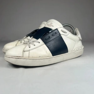 Valentino Garavani Open - Snygga Valentino Garavani Open sneakers i vitt skinn med bred marinblå läderdetalj över mitten. Är i hyfsat bra skick och använda fåtal gånger. Skorna är i storlek 41 och säljs för 1549kr. Nypris: 5500kr