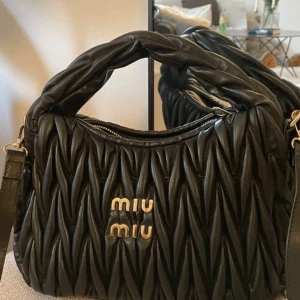 Svart quiltad handväska från Miu Miu - Supersnygg svart handväska från Miu Miu i quiltat skinn med guldiga detaljer och logga framtill. Väskan har en vadderad topphandtag, justerbar axelrem och dragkedja upptill. Perfekt för dig som vill ha en lyxig och trendig accessoar.