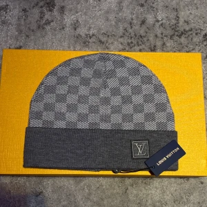 Grå rutig mössa från Louis Vuitton - Snygg grå mössa från Louis Vuitton med klassiskt rutigt mönster och vikt kant. Mössan har LV-logga framtill och är tillverkad i ett mjukt material som passar perfekt för kyliga dagar. En stilren accessoar som lyfter din vinterlook. Hör gärna av er vid frågor!