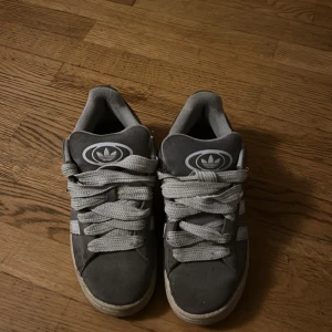 Adidas Campus grå sneakers mocka - Grå Adidas Campus sneakers i mocka med vita klassiska ränder på sidorna och breda vita skosnören. Snygg retrostil med gummisula och rund tå. Adidas logga på plösen och insidan. Perfekta för dig som gillar streetwear och skön stil.