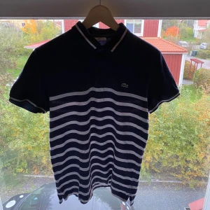 Randig pikétröja från Lacoste, marinblå - Säljer en klassisk marinblå och vit randig pikétröja från Lacoste med broderad krokodil på bröstet. Tröjan har korta ärmar, krage med knappar och sportig känsla. Perfekt för dig som gillar stilren och tidlös design.