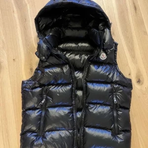 Svart dunväst från Moncler med huva - Snygg svart dunväst från Moncler med glansig finish, huva och två dragkedjefickor framtill. Klassisk Moncler-logga på bröstet och quiltad design som ger en cool och sportig vibe.
