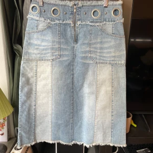 Ljusblå jeanskjol med metalldetaljer - Cool ljusblå jeanskjol från Sugar Jeans med råa kanter, stora metalldetaljer i midjan och dragkedja framtill. Kjolen har stora fickor både fram och bak samt en markerad sömstruktur som ger en unik look. Perfekt för dig som gillar statement-plagg.