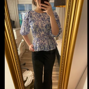 Blåvit blus  - Säljer en blåvit blus med mönster och puffiga trekvartsärmar. Blusen har markerad midja, volang nertill och öppen rygg med knytband upptill. Använd endast en gång!