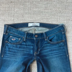 Blå bootcut jeans från Hollister - Snygga blå bootcut jeans från Hollister med klassisk femficksdesign och ljusa slitningar framtill. Jeansen har mellanblå tvätt, kontrastsömmar och broderad logga på myntfickan. Midjemåttet är 39 cm och innerbenslängden är 83 cm💓💓 Skriv gärna om ni har fler funderingar🥰(bilder från förra ägaren)
