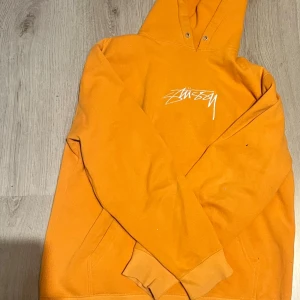 Orange hoodie från Stüssy - Säljer en orange hoodie från Stüssy med vit logga på bröstet. Tröjan har huva med snörhål, magficka och långa ärmar. Perfekt för dig som gillar streetwear och vill sticka ut med en färgstark look.
