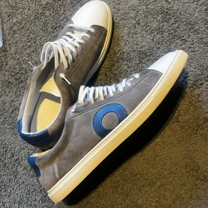 Grå mockasneakers från Oliver Cabell - Snygga sneakers från Oliver Cabell i grå mocka med vit tå och sula. Skorna har blå detaljer på hälen och sidan, samt vita skosnören. Klassisk låg modell med rund tå och platt sula, perfekt för en clean och stilren look.