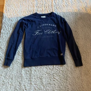 Mörkblå sweatshirt från J.Lindeberg - Snygg mörkblå sweatshirt från J.Lindeberg med vit text på bröstet. Tröjan har rund halsringning, långa ärmar och breda muddar vid ärmslut och nederkant. Perfekt för en stilren och avslappnad look.