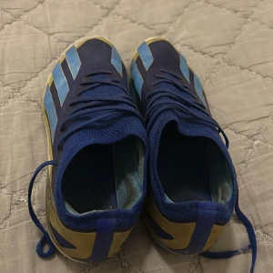 Adidas Messi blå fotbollsskor elite - Säljer ett par Adidas Messi fotbollsskor i blått med ljusblå och guldiga detaljer. Skorna har snörning, låg profil och mönstrad sula med dobbar för bra grepp på planen. Ovandelen är i syntetmaterial och designen är sportig och modern elite.