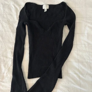 Svart ribbad långärmad topp - Säljer en svart ribbad långärmad topp med markerad byst och figurnära passform. Toppen har en djupare urringning och är tillverkad i stretchigt material