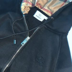 Svart Burberry vindjacka med huva - Svart vindjacka från Burberry i 100% bomull med klassiskt rutigt foder i huvan. Jackan har dragkedja framtill, snörning vid huvan och diskret broderad logga på bröstet. Perfekt för dig som vill ha en stilren och ikonisk look.