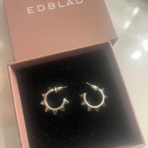 Silvriga örhängen med nitar från Edblad - Coola silvriga örhängen från Edblad i form av små hoops med spetsiga nitar runt om. Perfekta för dig som vill ha en edgy och unik detalj till din stil. Kommer i originalask.