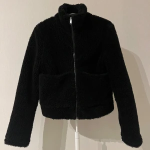 Svart teddyjacka med dragkedja - Jag säljer en svart teddyjacka med hög krage och hel dragkedja framtill. Jackan har två stora fickor på framsidan och den är lite kortare i modellen. Varan har inga tecken på användning.