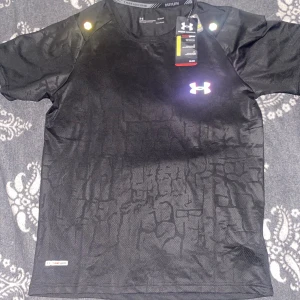Svart Under Armour Reflective t-shirt - Svart Under Armour t-shirt i funktionsmaterial med diskret mönster och reflekterande logga på bröstet. Kortärmad modell med rund hals och HeatGear-teknologi som håller dig sval och torr. Perfekt för löpning eller träning.