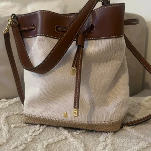 Beige och brun axelväska från Ralph Lauren - Snygg axelväska i beige canvas med bruna skinn-detaljer och flätad botten. Väskan har justerbar axelrem, dragskostängning med guldfärgade RL-detaljer och rymligt innerfack. Perfekt för dig som gillar klassisk och stilren design.