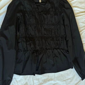 Svart blus med v-ringning från Åhléns - Svart blus i glansigt material från Åhléns med v-ringning och långa ärmar. Blusen har smockdetaljer över hela livet och lätt volang nertill. Perfekt för dig som gillar en stilren men ändå cool look.