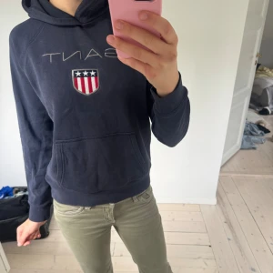 Mörkblå hoodie från Gant - Säljer en mörkblå hoodie från Gant i kvinno modell, med broderad logga och amerikanskt inspirerat märke på bröstet. Tröjan har huva, känguruficka och långa ärmar. Perfekt för en chill och sportig look.
